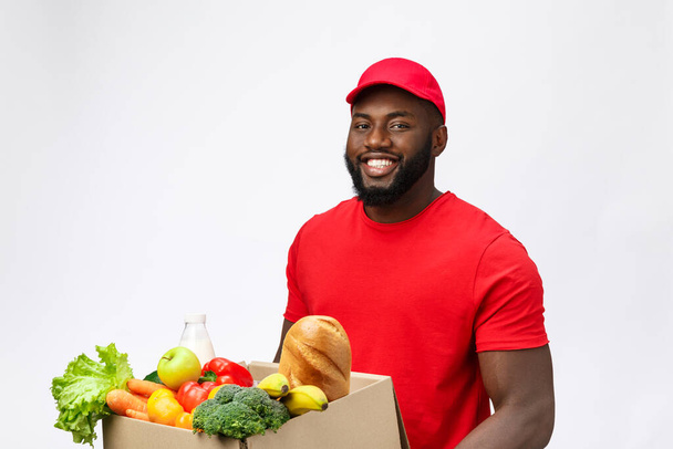 Delivery Concept - Handsome African American delivery man carrying package box of grocery food and drink from store. Isolated on Grey studio Background. Copy Space - Photo, Image