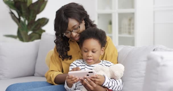 Αφροαμερικανή μητέρα με γυαλιά που δείχνει κάτι στη μικρή χαριτωμένη κόρη της στην οθόνη του smartphone. Η μαμά με το μικρό παιδί να χτυπάει, να κυλάει και να παίζει στο κινητό. Διδασκαλία παιδιού με κινητό. - Πλάνα, βίντεο