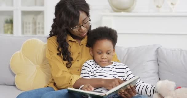 Νεαρή όμορφη Αφροαμερικανή γυναίκα σε γυαλιά διαβάζοντας βιβλίο με μικρή χαριτωμένη κόρη στον καναπέ στο σπίτι. Μικρό παιδί γυρίζοντας σελίδες παραμυθιού με φωτογραφίες με τη μητέρα στον καναπέ. Babysitter. - Πλάνα, βίντεο