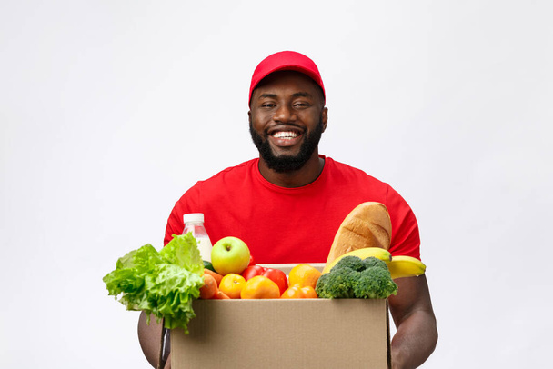 Concepto de entrega: el guapo repartidor afroamericano lleva una caja de alimentos y bebidas de la tienda. Aislado en Grey studio Background. Copiar espacio
 - Foto, Imagen