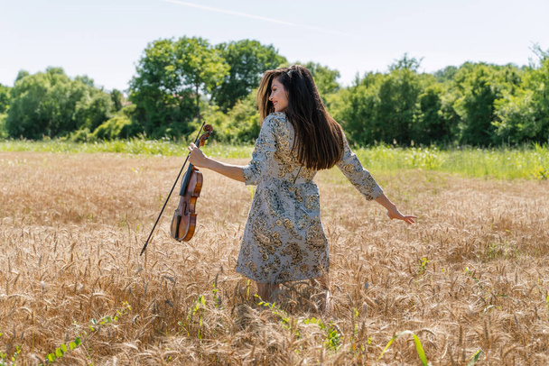 Portrait of adult caucasian female in the wheat field holding violin - Woman in summer day dancing alone outdoor full length back view - freedom nature solitude mother nature concept - Photo, image