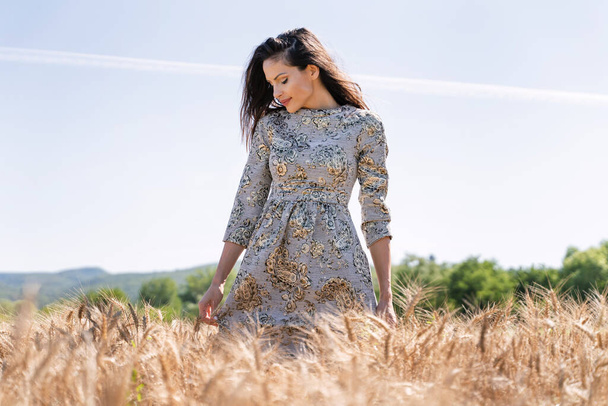 Front view on beautiful caucasian woman wearing dress in the crops - Pretty female adult standing in wheat field looking down in summer day - identity solitude mother nature concept - Photo, Image