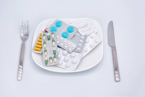Diet.  Different  pills, medicines, drugs on a white square shaped plate with fork and knife around isolated on white background. Taking pills instead of food, unhealthy sick lifestyle concept - Photo, image