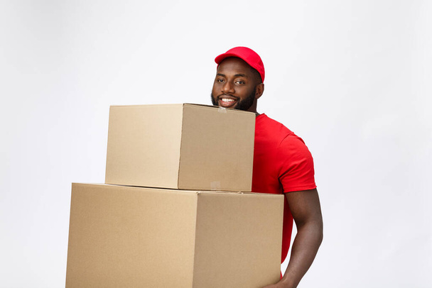 Lieferkonzept - Seitenansicht Porträt eines glücklichen afroamerikanischen Zustellers in rotem Tuch, der ein Karton-Paket hält. Isoliert auf grauem Hintergrund. Kopierraum - Foto, Bild
