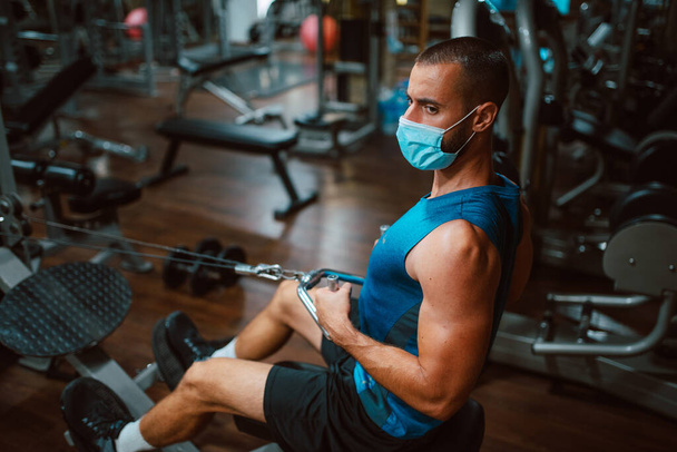 Jeune homme de fitness musculaire avec masque médical faisant un exercice de poids lourd pour le dos et le bras sur la machine dans la gymnase. - Photo, image