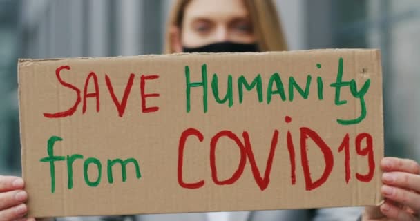 Закрыть картонный стол словами Save Humanity from Covid 19 в руках белой молодой женщины в маске. Расплывчатая женская доска. Одинокий протест на открытом воздухе. - Кадры, видео