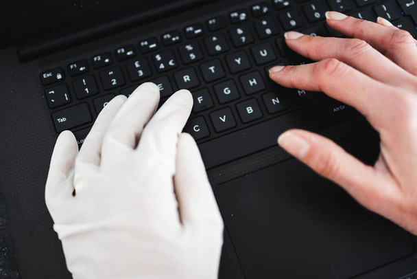 staying safe after the covid-19 virus pandemic outbreak, hand typing on shared computer keyboard at work wearing disposable glove to avoid contact with potentially infected surfaces and other hand without glove - 写真・画像