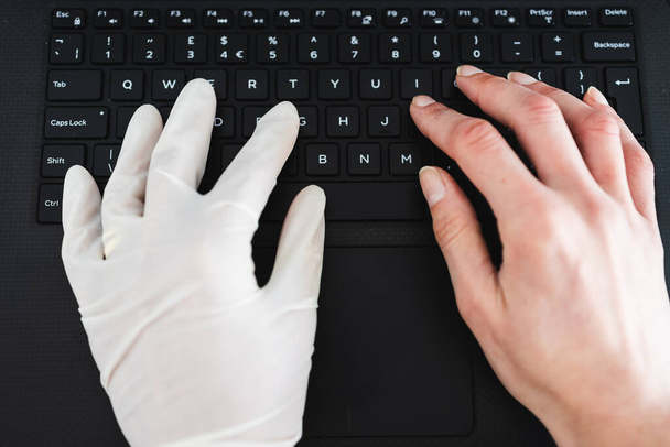 staying safe after the covid-19 virus pandemic outbreak, hand typing on shared computer keyboard at work wearing disposable glove to avoid contact with potentially infected surfaces and other hand without glove - Фото, зображення