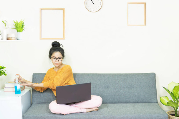 people and leisure concept - asian young woman with laptop working on sofa, Wash her hands with an alcohol gel while watching the screen, resting on weekends at home under quarantine - Zdjęcie, obraz