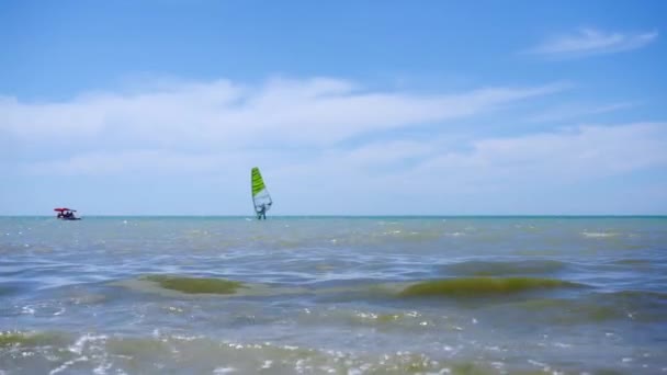 Άποψη της ακτής της θάλασσας σε μια καθαρή ηλιόλουστη μέρα. Μια σιλουέτα ενός windsurfing άνθρωπος είναι ορατό στο βάθος. Σκηνή ανάπαυσης δίπλα στη θάλασσα ή στον ωκεανό. - Πλάνα, βίντεο
