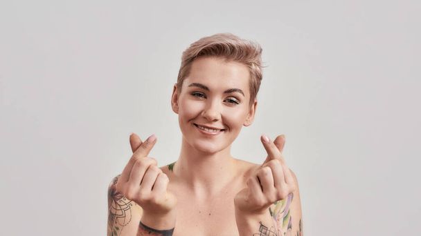 Spread Love. Portrait of cheerful half naked tattooed woman with pierced nose and short hair smiling at camera, showing Korean heart sign isolated over light background - Foto, Bild