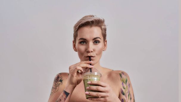 Smoothie. Attractive tattooed woman with pierced nose and short hair looking at camera, while drinking green detox smoothie isolated over grey background - Photo, image