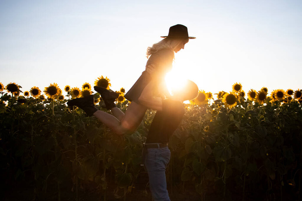 Kaunis nuori pari suutelee auringonlaskun aikaan auringonkukkapellolla. Blondi mustassa hatussa ja julma parrakas mies. Elämäntapa, rakkaus ja rentoutuminen käsite - Valokuva, kuva