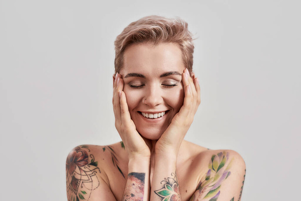 Close up portrait of a young attractive woman with short hair, perfect skin and makeup smiling with closed eyes, touching face isolated over light background - Photo, Image