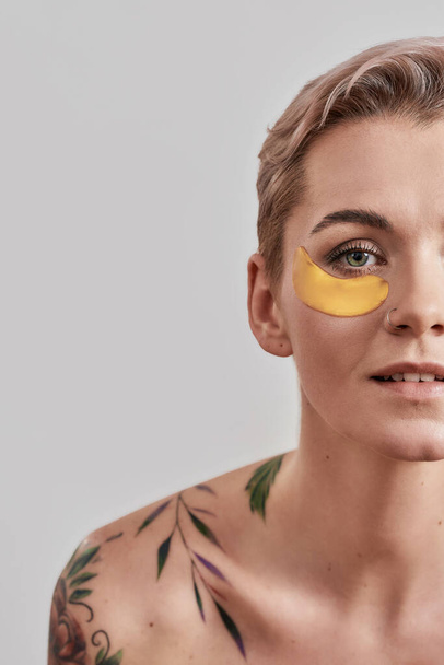 Cropped portrait of half naked tattooed woman with short hair looking up, caring for her face, wearing under eye patches for dark circles isolated over light background - Photo, image