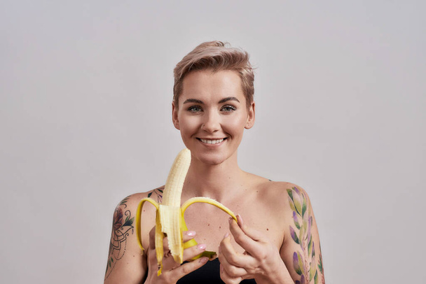 Your protein supplement. Attractive tattooed woman with pierced nose and short hair smiling at camera while peeling banana standing isolated over grey background - Foto, afbeelding