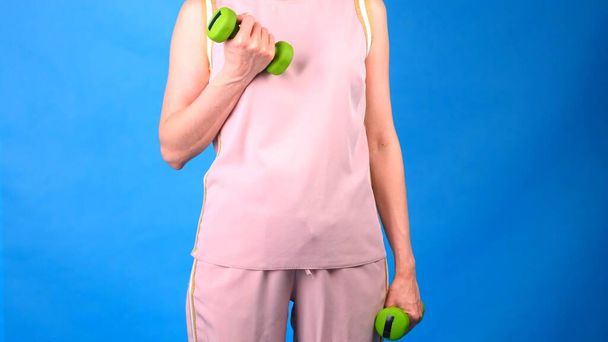 fat Female in a pink sports suit with dumbbells in hands does exercises on a blue background. The concept of sports, diet and weight loss. - Photo, Image