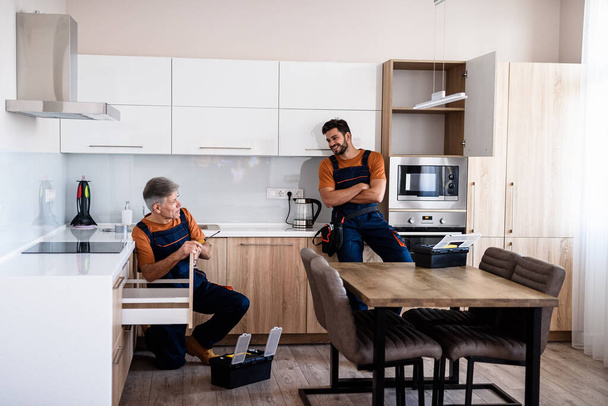 Full length shot of two handymen, workers in uniform talking while assembling kitchen cabinet using screwdriver indoors. Furniture repair and assembly concept - Foto, immagini