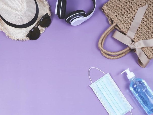 COVID-19 Prävention, Reisen und neues normales Konzept, Draufsicht auf gewebte Tasche mit Chirurgenmaske und Desinfektionsgel und Urlaubs-Accessoires für Frauen auf lila Hintergrund. - Foto, Bild