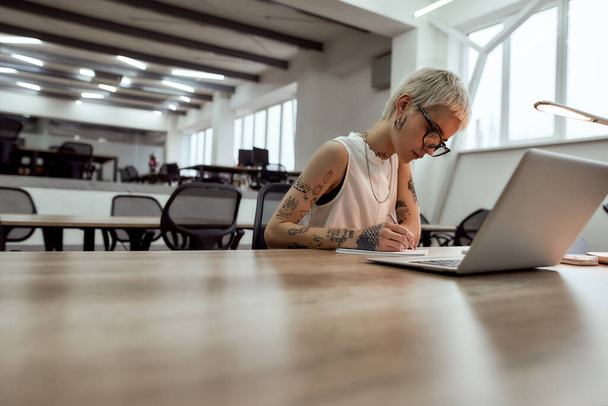 Young, beautiful tattooed business lady in eyewear making some notes while sitting alone in the modern office - Fotó, kép