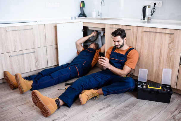 Young repairman, worker in uniform sitting on the floor, using smartphone while his experienced colleague fixing sink pipe indoors. Repair service, new generation concept - Zdjęcie, obraz
