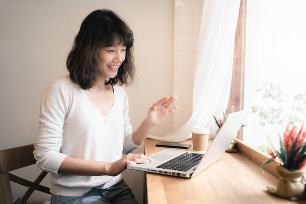 Jovem mulher asiática trabalhando e videoconferência com computador portátil. Uma mulher feliz com um rosto sorridente a trabalhar em casa. Uma chávena de café na mesa. Trabalho a partir do conceito de casa. Bangkok, Tailândia
 - Foto, Imagem