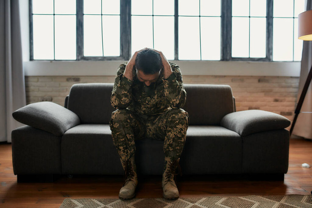 Constant pain. Full length shot of middle aged military man holding his head in pain and depression sitting on the couch. Soldier suffering from psychological trauma. PTSD concept - Φωτογραφία, εικόνα