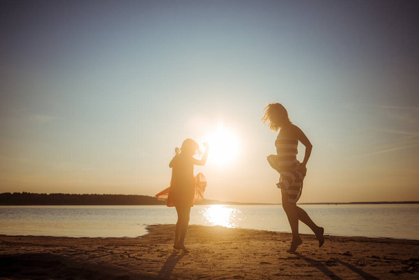 Счастливые мама и дочь в платьях прыгают и танцуют на пляже во время заката. Хорошие отношения двух поколений. Веселое семейное времяпрепровождение на природе. Продвижение здоровья через игры и мероприятия на открытом воздухе. - Фото, изображение