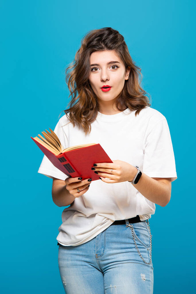 Young student, teenage girl with red lips holds a book in hands, isolated on a blue background. Surprised face - Фото, изображение