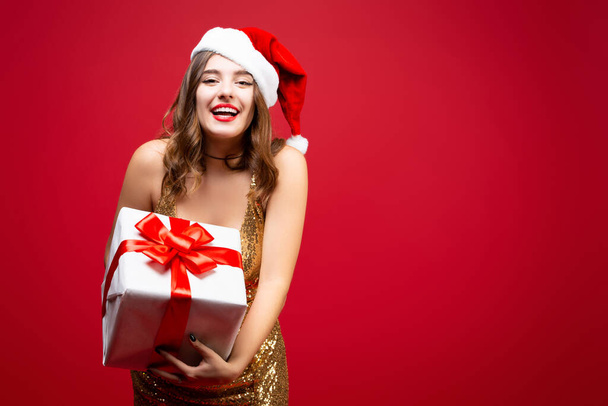 Beautiful sexy Santa girl in gold elegant evening dress and Christmas hat with a New Year gift in hands. Emotions happiness and delight. Smiling face - Foto, imagen