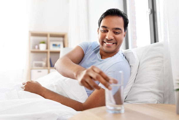 happy indian man drinking water lying in bed - Foto, afbeelding