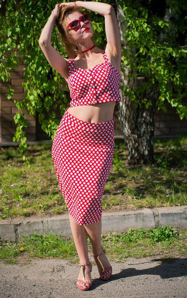 Una juguetona chica dulce y sexy americana en un traje pin up con lunares con labios rojos y gafas de sol. Una mujer en traje vintage pasea por una pequeña ciudad
 - Foto, imagen
