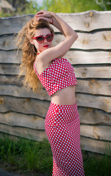 A playful American sweet and sexy girl in a pin up suit with polka dots with red lips and sunglasses. A woman in a vintage suit strolls through a small town - Photo, image