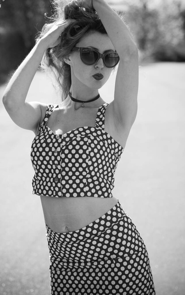 A playful American sweet and sexy girl in a pin up suit with polka dots with red lips and sunglasses. A woman in a vintage suit strolls through a small town - Fotoğraf, Görsel