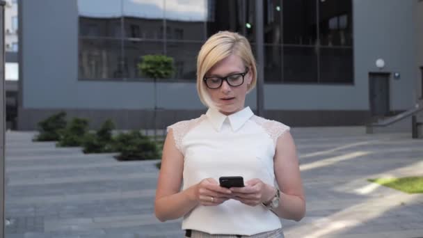 Mooi blond in bril business dame gaat naar buiten maakt gebruik van een smartphone om een zakelijke bijeenkomst te regelen ze is gekleed in een business suit en heeft prachtig vormgegeven haar en een stijlvol horloge. - Video