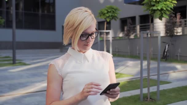 Ελκυστική επιχειρηματίας με γυαλιά πηγαίνει σε εξωτερικούς χώρους χρησιμοποιεί ένα κινητό τηλέφωνο για να αγοράσει κάτι που είναι ντυμένη με ένα επιχειρηματικό κοστούμι και έχει όμορφα στυλ μαλλιά και ένα κομψό ρολόι. - Πλάνα, βίντεο