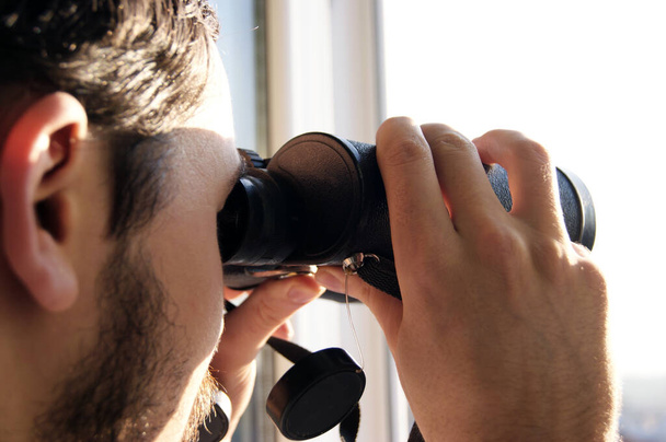 Jongeman die door een glazen raam kijkt met een verrekijker terwijl hij iets in de verte bekijkt. - Foto, afbeelding