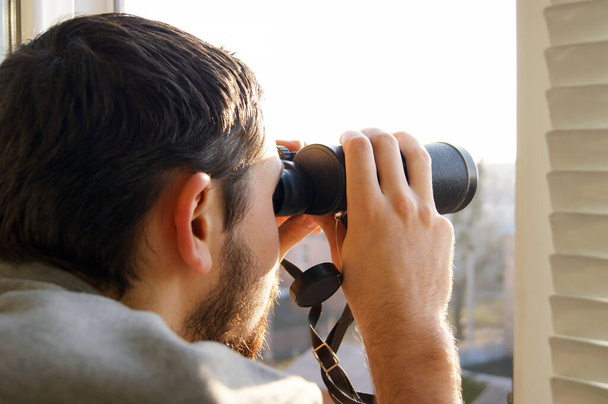Young man standing looking through a glass window with binoculars as he watches something in the distance. - Photo, Image