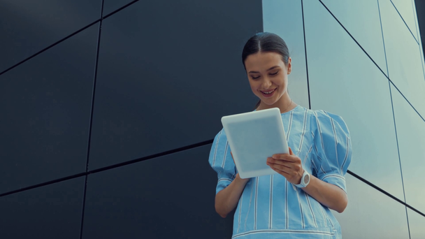 nízký úhel pohledu šťastné ženy pomocí digitálního tabletu při stání v blízkosti budovy - Záběry, video