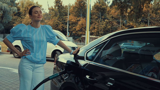 attraktive Frau, die mit der Hand auf der Hüfte steht, während sie Auto an Tankstelle tankt - Filmmaterial, Video