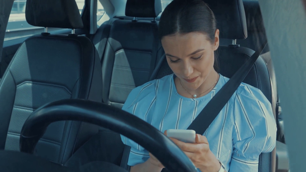 smiling woman chatting on smartphone while sitting in car on drivers seat  - Felvétel, videó
