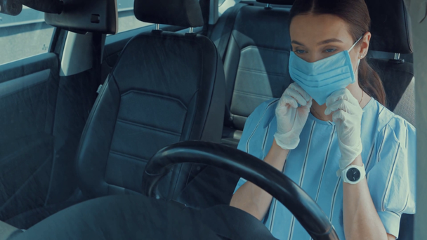 młoda kobieta w lateksowych rękawiczkach zakładająca maskę medyczną podczas siedzenia w samochodzie - Materiał filmowy, wideo