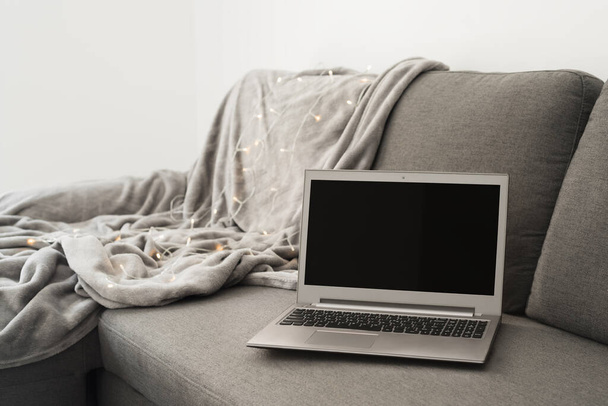 Φορητός υπολογιστής με κενή μαύρη οθόνη στέκεται σε γκρι καναπέ στο σαλόνι, ζεστή κουβέρτα και διακοσμητικά φώτα ως φόντο - Φωτογραφία, εικόνα