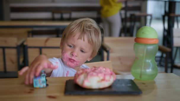 desserts, kleine mannelijke kind met zoete room op zijn gezicht wordt gespeeld door de machine na snack met lekkere donut zitten aan tafel in cafe - Video