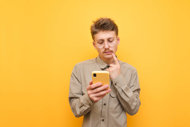 Thoughtful young man with mustache and glasses uses a smartphone against a yellow background,looks at the screen and thinks,wearing a shirt.Funny nerd with a serious face is using the phone. Isolated - Photo, image