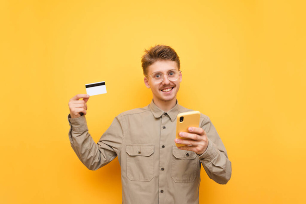 Een jongeman in bril en shirt staat op een gele achtergrond met een smartphone en een bankkaart in zijn hand, kijkt in de camera en lacht. Nerd met krediet en telefoon geïsoleerd. Winkelen Internet. - Foto, afbeelding