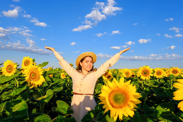 glimlachende vrouw in strohoed in bloeiende zonnebloemenveld met uitgestrekte armen  - Foto, afbeelding