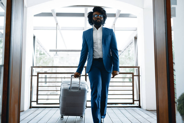 Black man in formal suit with packed suitcase entering hotel door - Фото, изображение