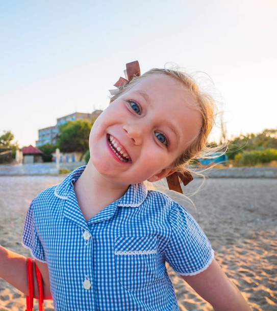 Портрет красивой девушки в голубом клетчатом летнем платье с блондинистыми волосами крупным планом. Радостный милый ребенок улыбается на фоне природы. Позитивный эмоционально смеющийся ребенок на пляже с морским песком. - Фото, изображение