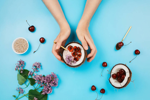 Flatlay with woman's hand holding half of coconut with plant-based yogurt bowl with fresh cherry and chia seeds on side on blue background - Foto, Imagen